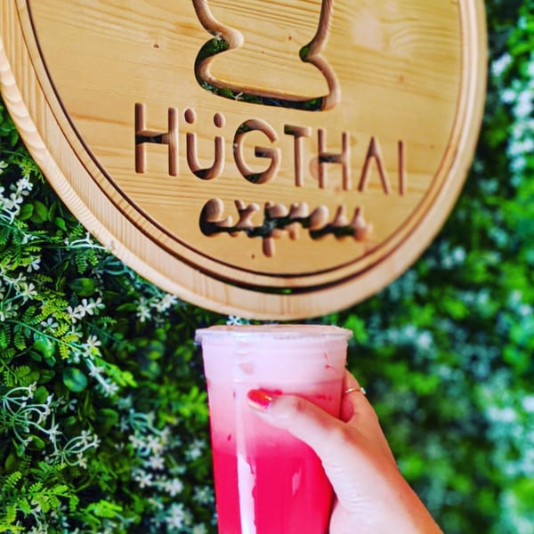Hand holding a vibrant pink drink in front of a Hug Thai Express wooden sign and green foliage.